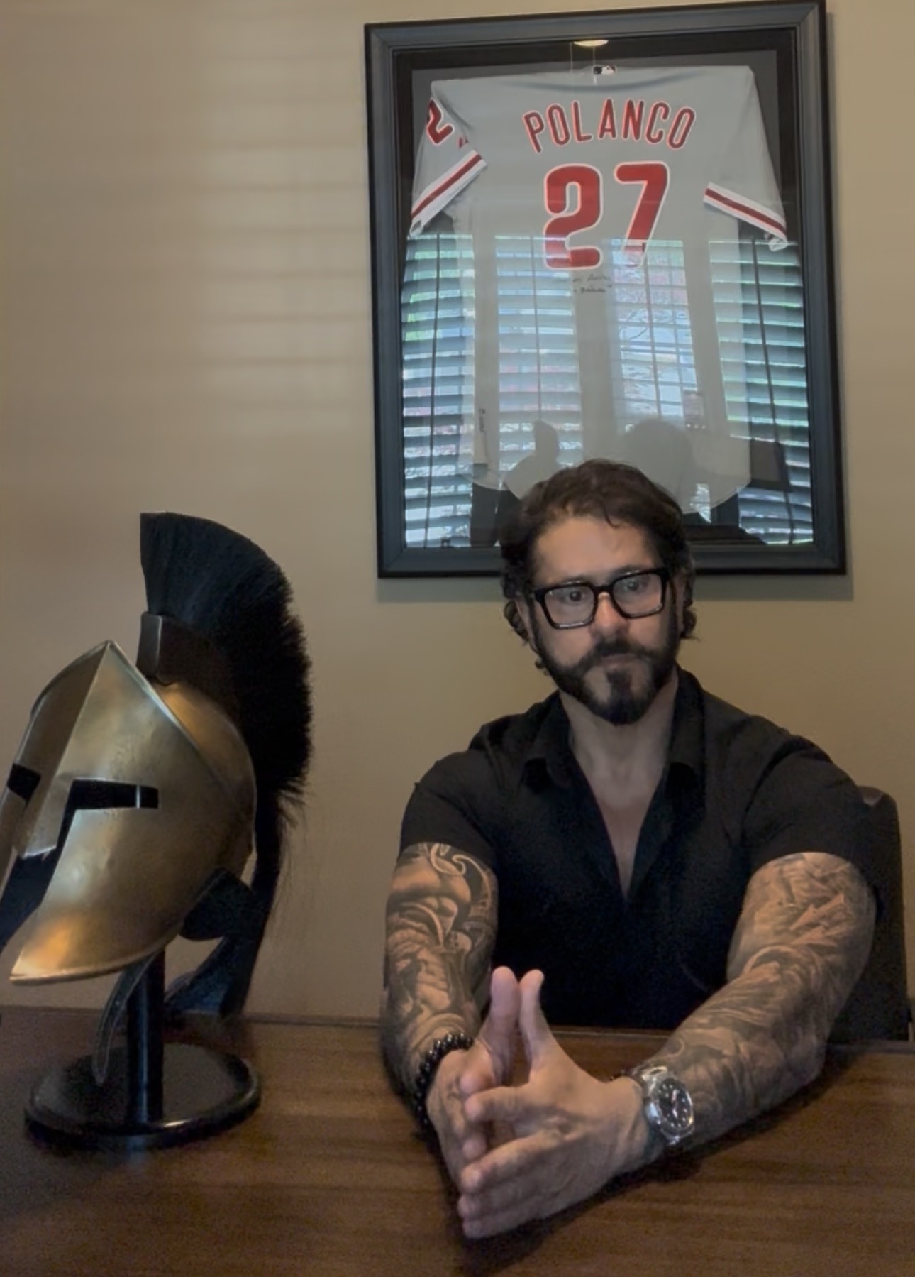 Man with tattoos sits at a table beside a bronze Spartan helmet. A framed "Polanco 27" jersey hangs on the wall. The scene is calm and contemplative.
