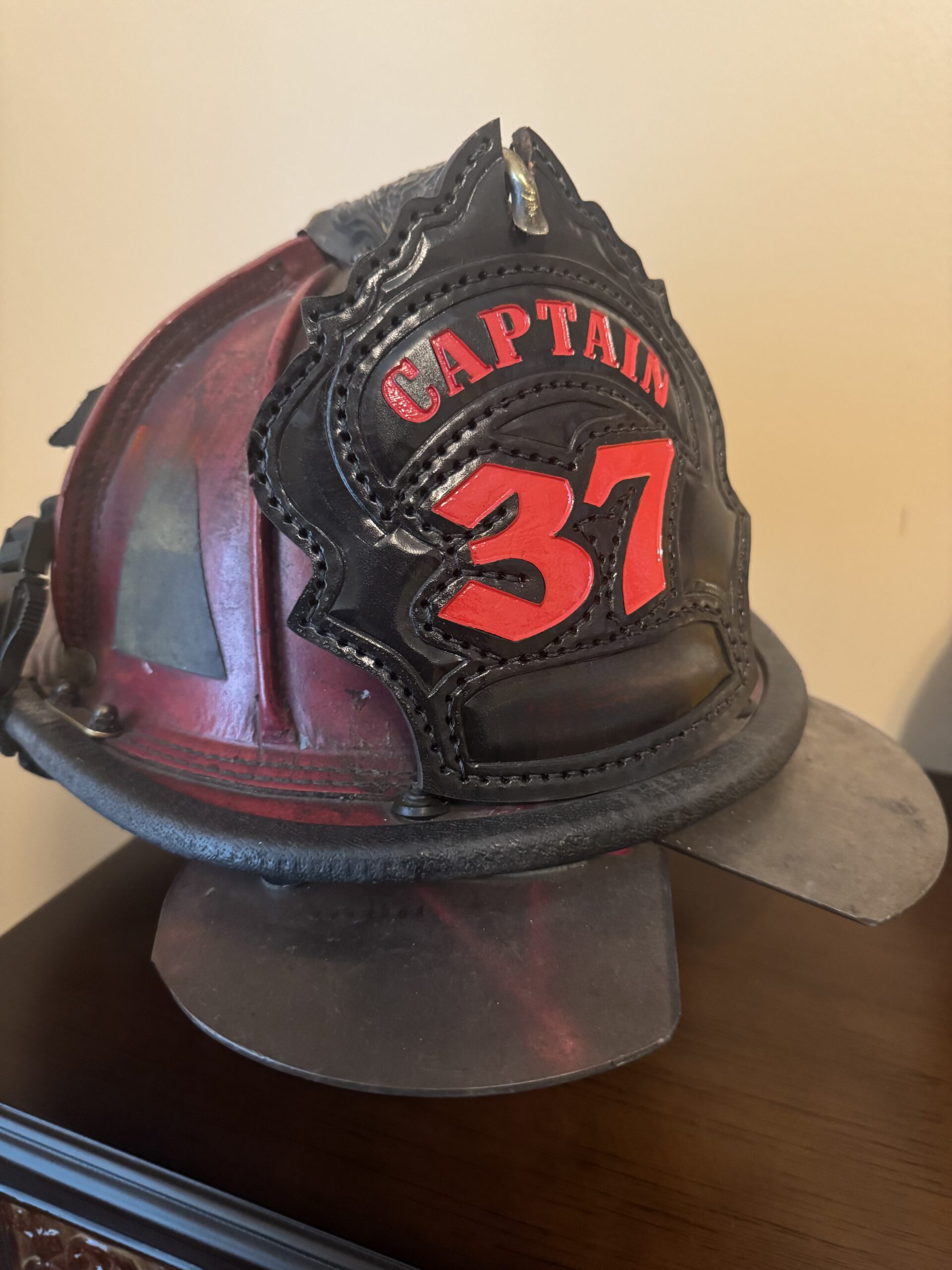 A worn firefighter's helmet sits on a wooden surface. It is red with a black front shield displaying "Captain 37" in bold, red lettering. The helmet conveys a sense of bravery and leadership.