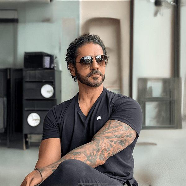 Man with curly hair and beard, wearing sunglasses and a black t-shirt, showcases a detailed tattoo sleeve on his crossed arms, exuding a confident vibe.