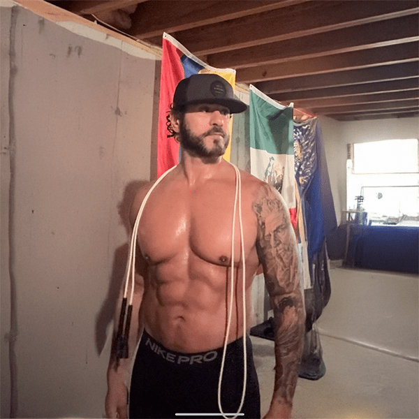 A muscular man wearing a black cap and shorts stands shirtless with a jump rope around his neck in a room with visible flags and exposed ceiling beams.