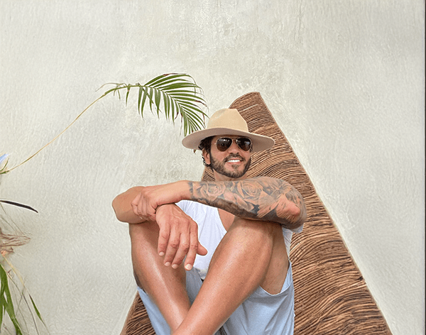 Man with a beard smiling, wearing a beige hat and sunglasses, sits against a textured wall. A tattooed arm is visible, and a leafy plant adds a relaxed vibe.