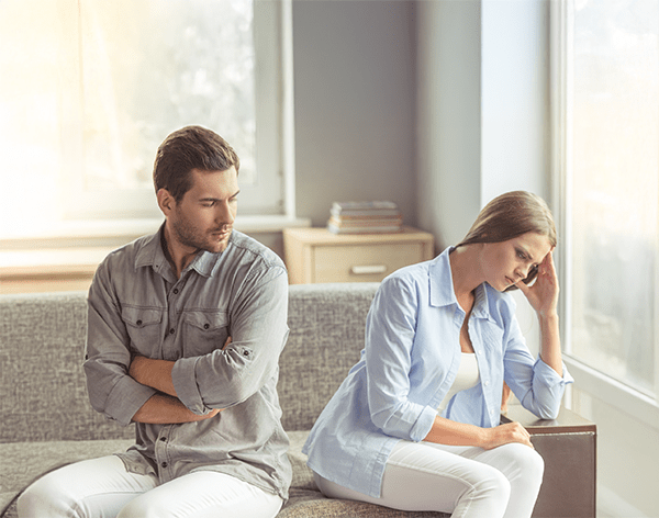 A man and woman sit on a couch, facing away from each other. The man looks thoughtful with arms crossed, while the woman rests her head on her hand, appearing upset. Natural light filters through a window, casting a somber ambiance.